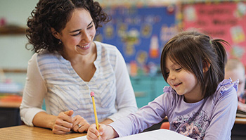 Profesora sonriendo mientras ve a una alumna hablar mientras escribe