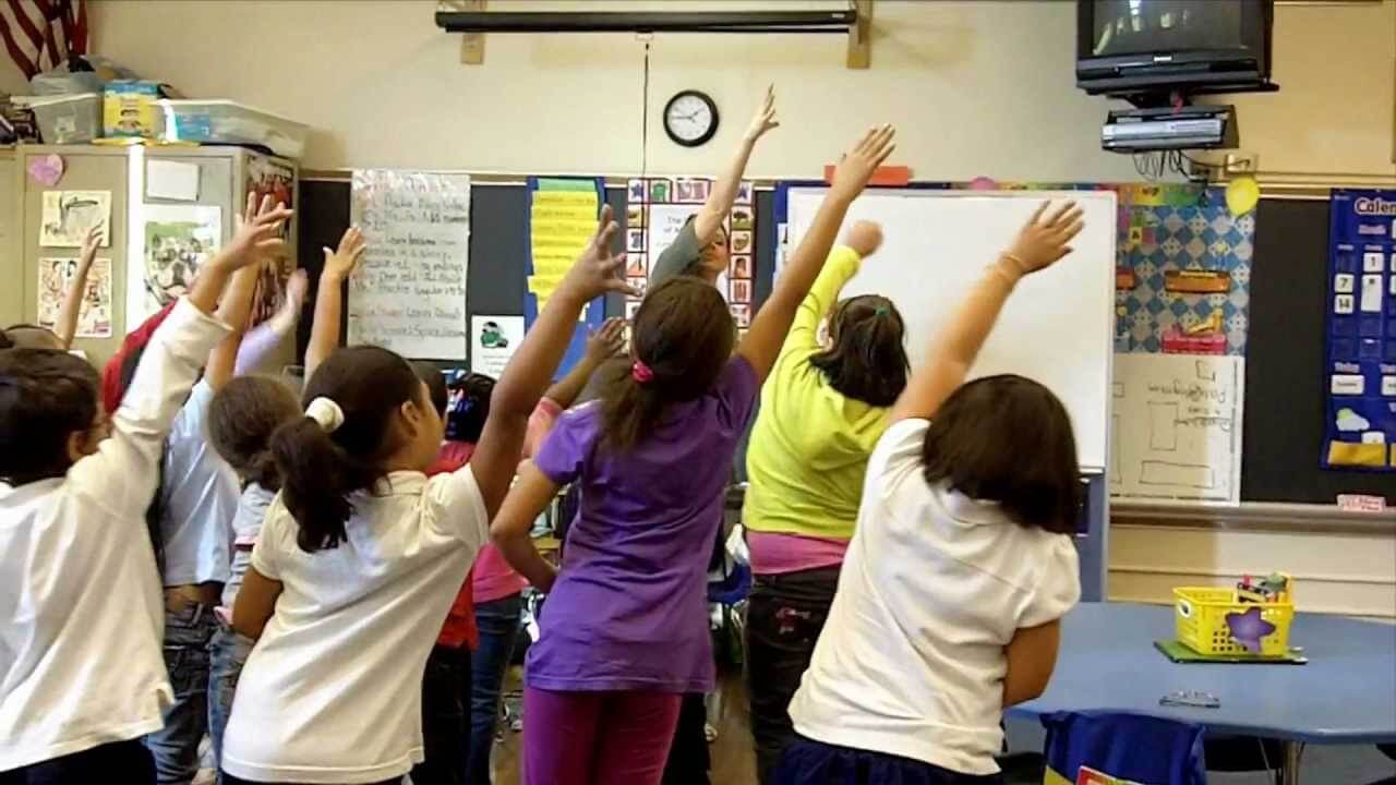 Niños en un salón de clase estirando los brazos hacia arriba y los costados durante clase para relajarse